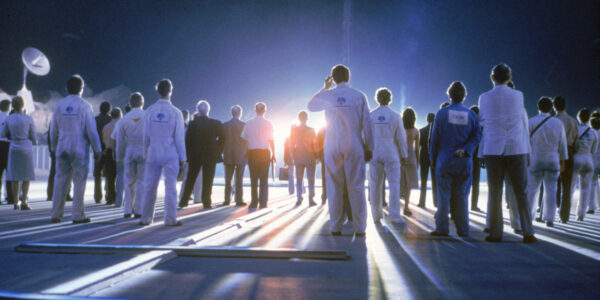 A group of people shot from behind stand on a runway as a bright orange light casts their long shadows on the ground