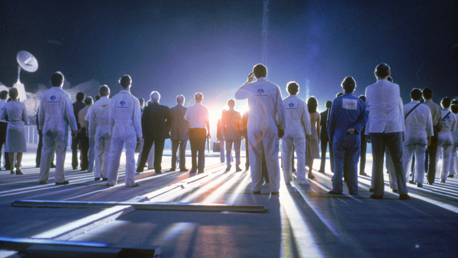A group of people shot from behind stand on a runway as a bright orange light casts their long shadows on the ground