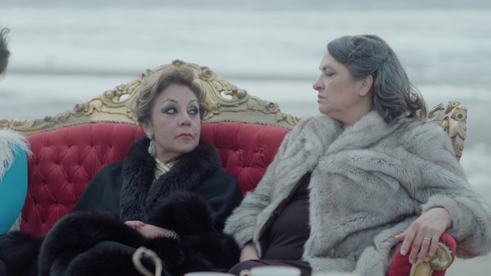 Film still- Before I Forget by Mariam Mekiwi Two women in fur coats sit on a red sofa outdoors and look at each other
