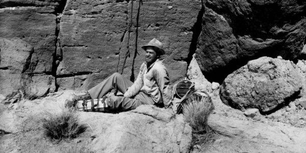 A man in a hat since in the desert by the side of a rocky mountain wall, holding his injured leg.