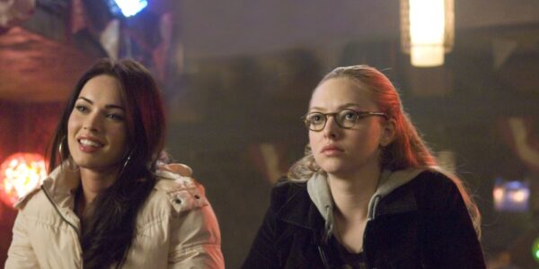 Two young women sit at a bar, one blonde, one brunette, the blonde isn't smiling, the brunette is, as they look off camera