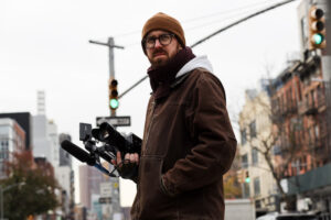 A man in glasses holds a video camera as he stands on a New York City street