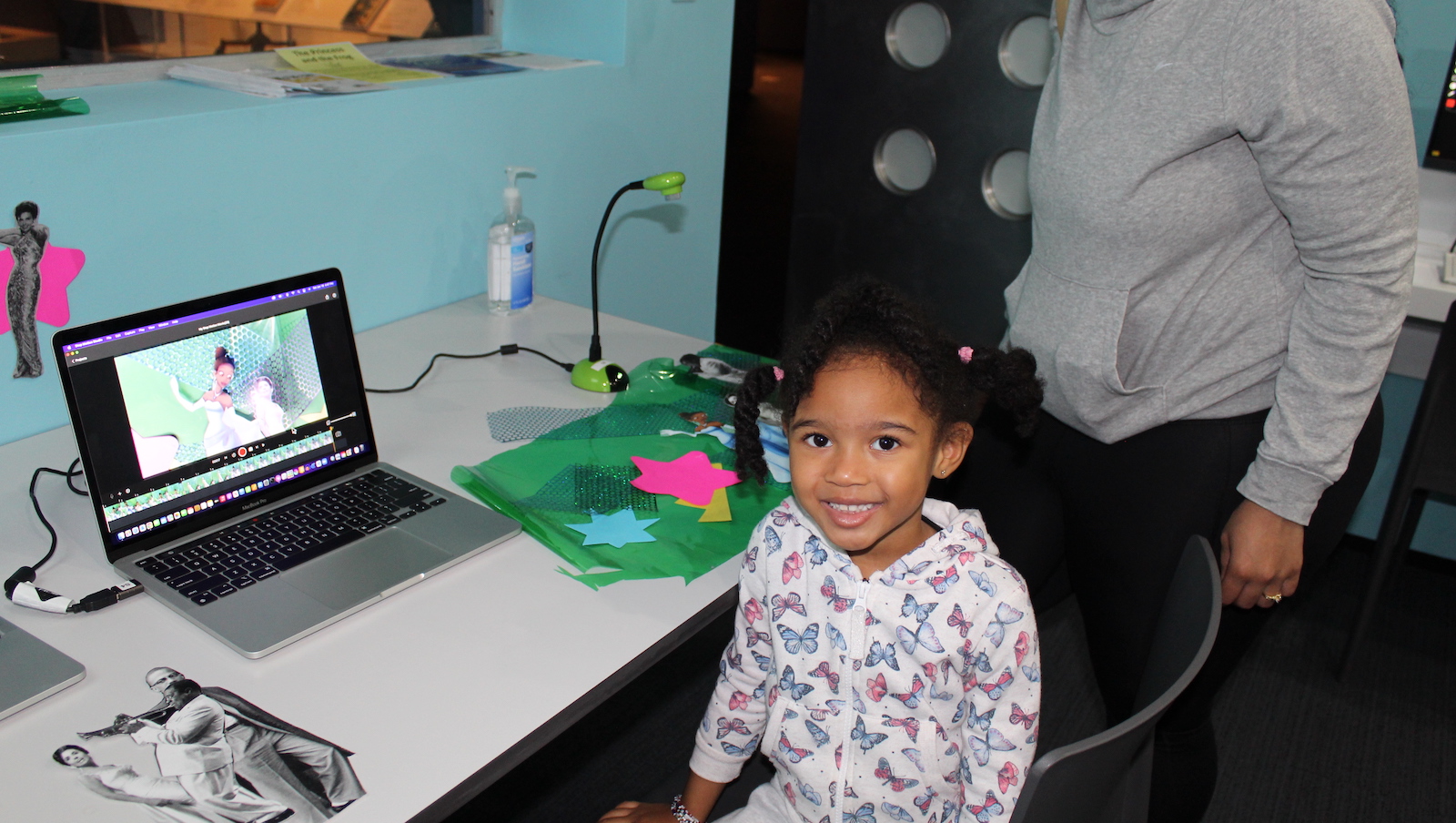 A little girl sits in front of a laptop computer with an animated princess on the screen, and she looks at the camera smiling