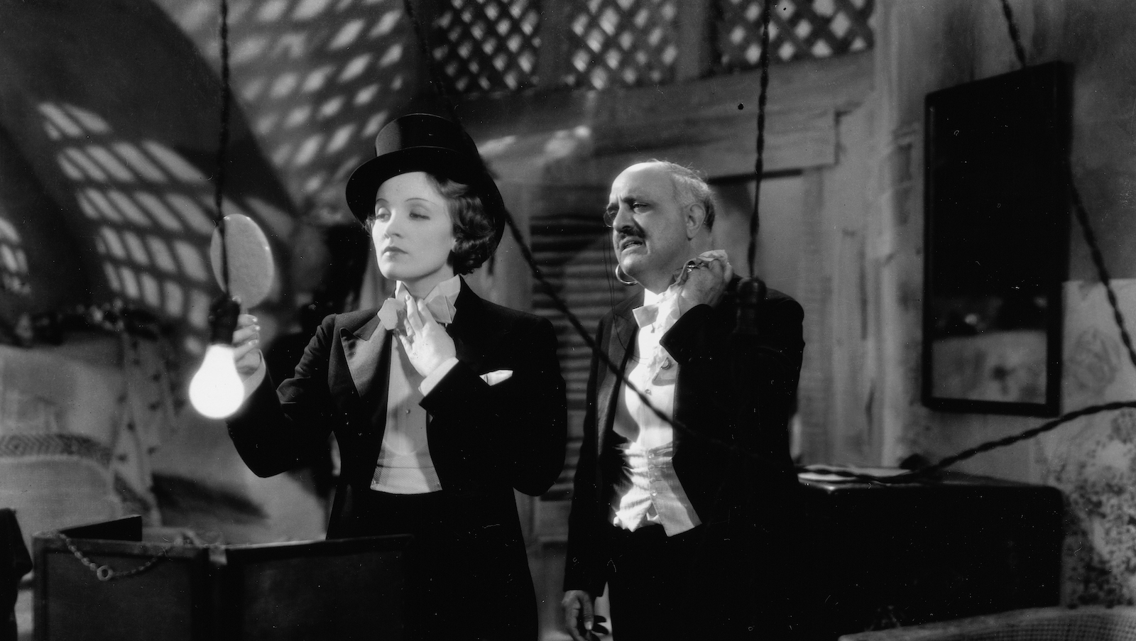 a woman in a tuxedo backstage ties her necktie and looks in a hand mirror, next to a mustachioed man