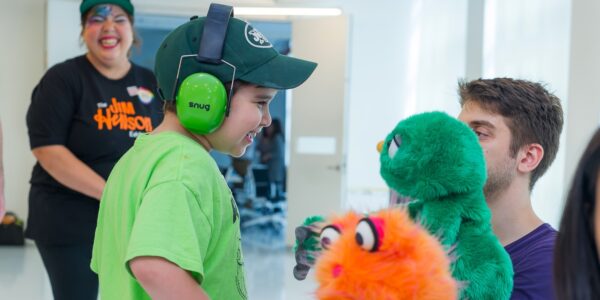 A little boy with green headphones and a green shirt interacts with a green puppet ...another orange puppet is in foreground.