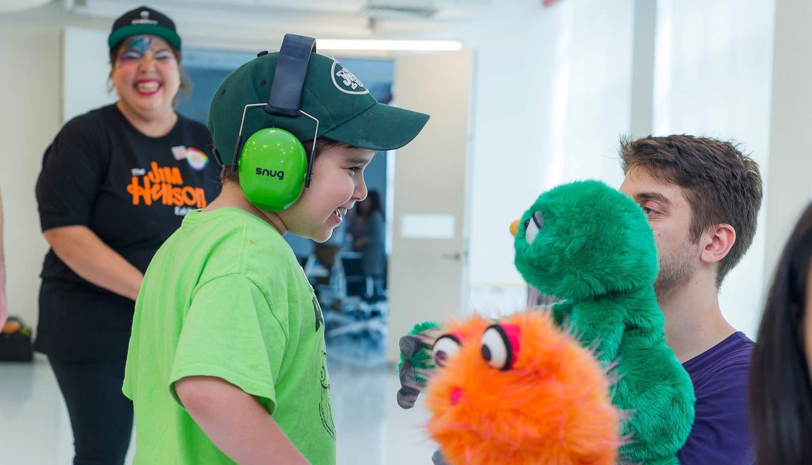 A little boy with green headphones and a green shirt interacts with a green puppet ...another orange puppet is in foreground.