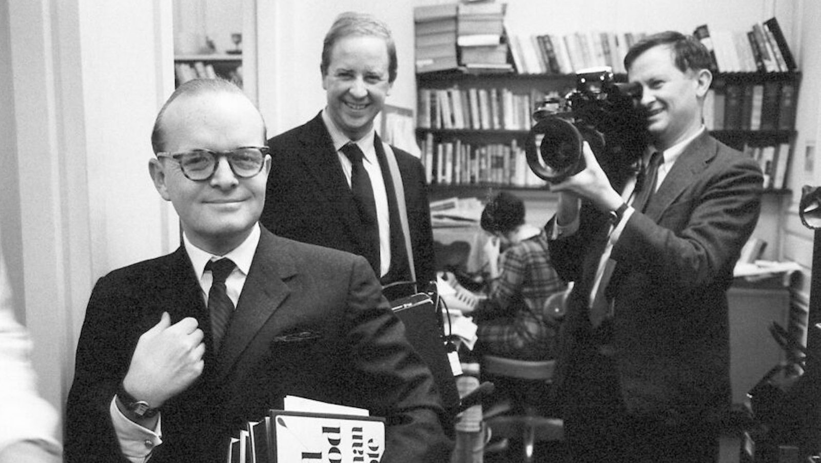 A man in a sharp suit and spectacles holds his lapel as two men behind him film him with a camera