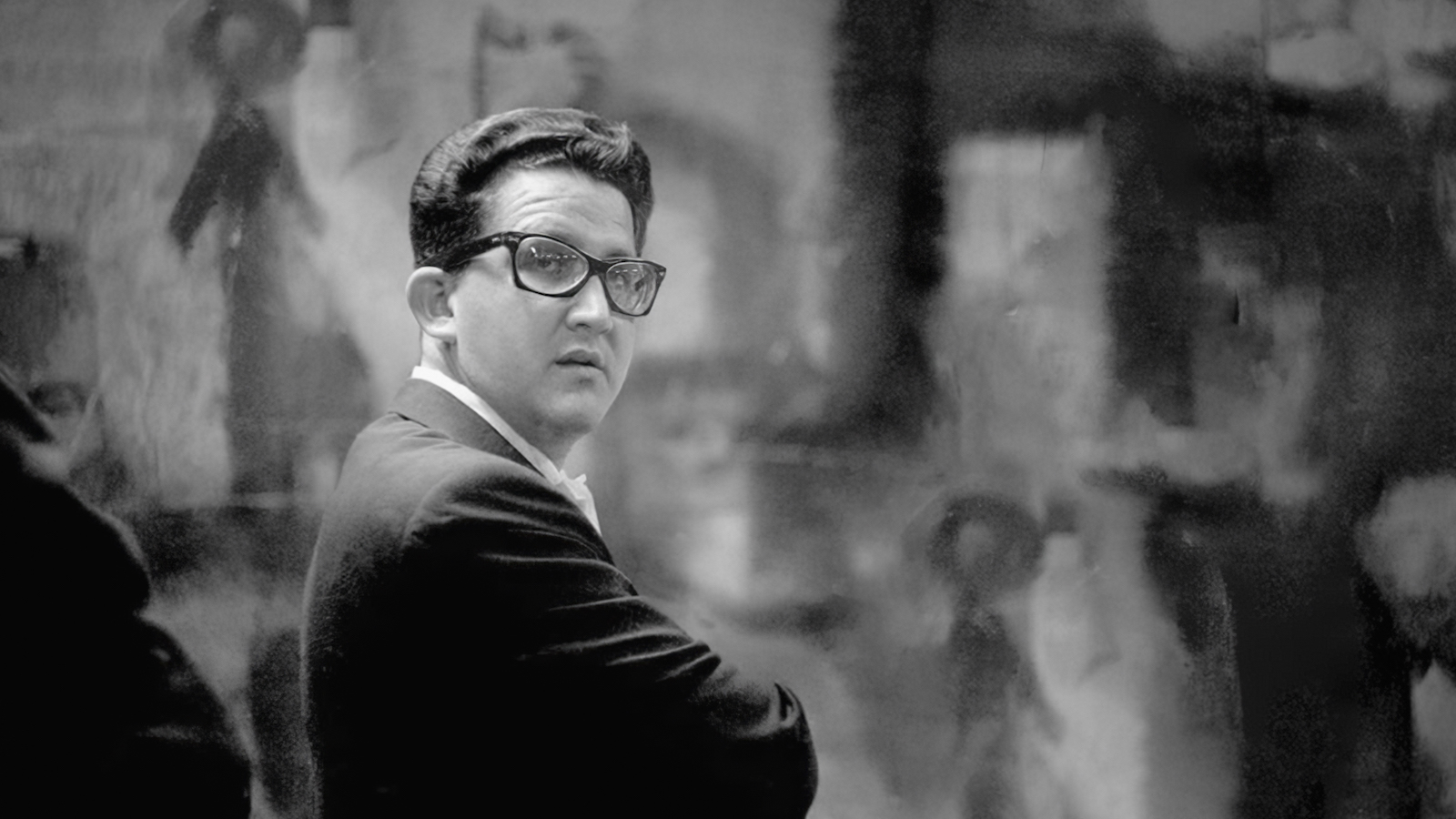 A black and white photo of a man in glasses and suit jacket standing with his arms crossed and a serious expression.