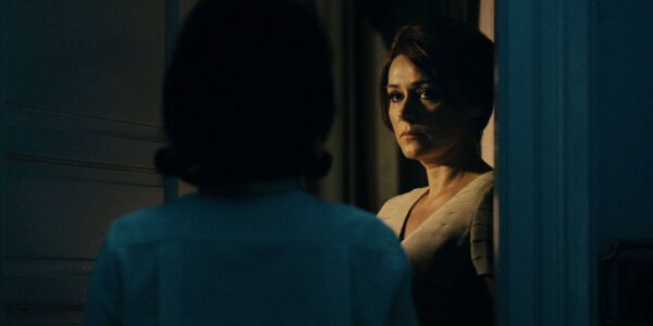 A woman looks at a woman—whom we see from behind—in a dark hallway