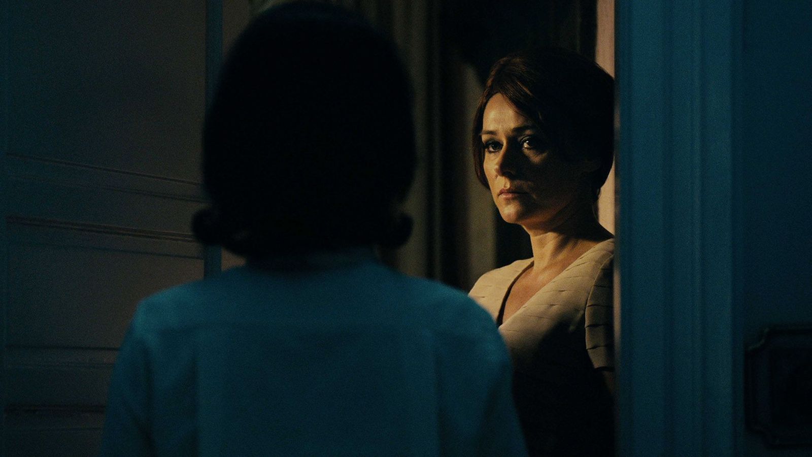 A woman looks at a woman—whom we see from behind—in a dark hallway