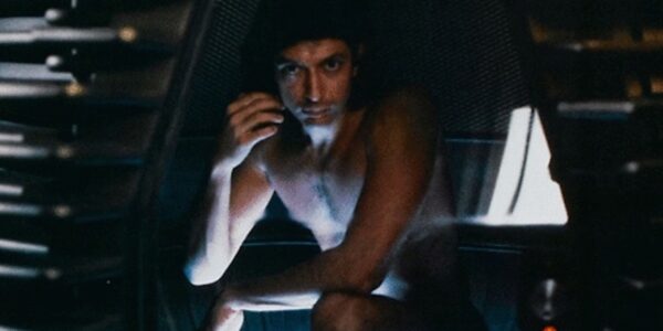 A naked man crouches in a futuristic machine pod, intensely looking at camera with one hand near his cheek