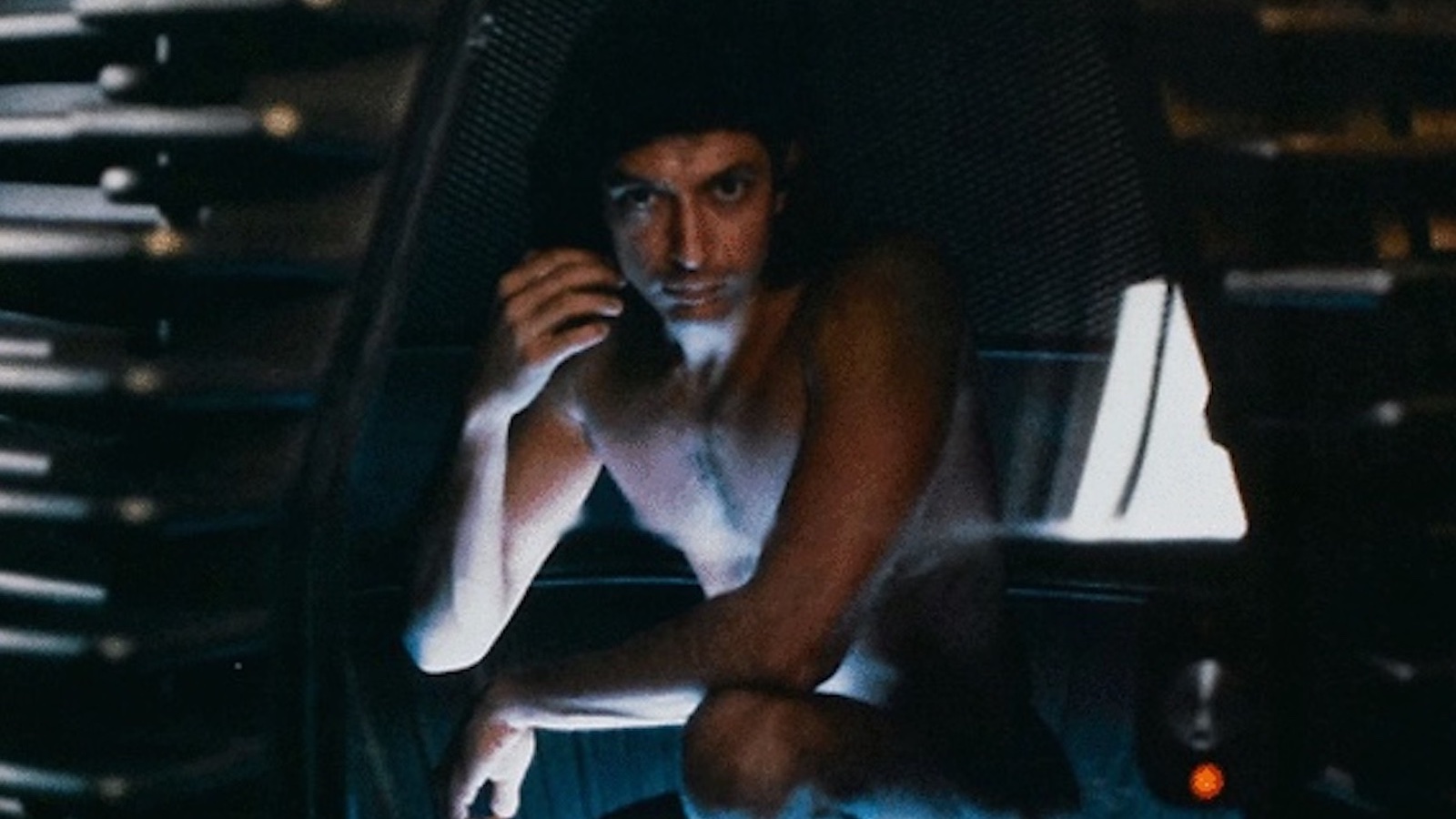 A naked man crouches in a futuristic machine pod, intensely looking at camera with one hand near his cheek