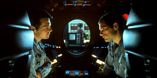 Two astronauts in profile sit in an enclosed pod and discuss with an oval shaped window behind them showing a computer's red light pointed in the direction of the camera
