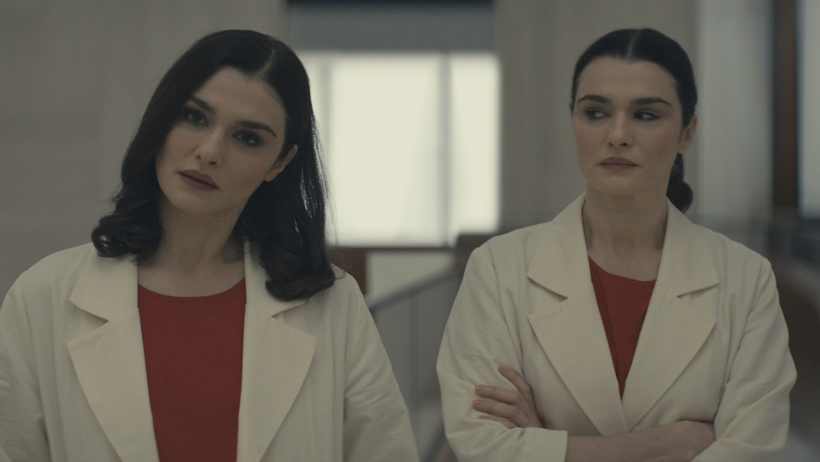 Two women in white doctor smocks and red shirts, played by the same dark-haired actress, stand side by side, one on the left looking off camera, the one on the right looking at the one on the left with her arms folded.