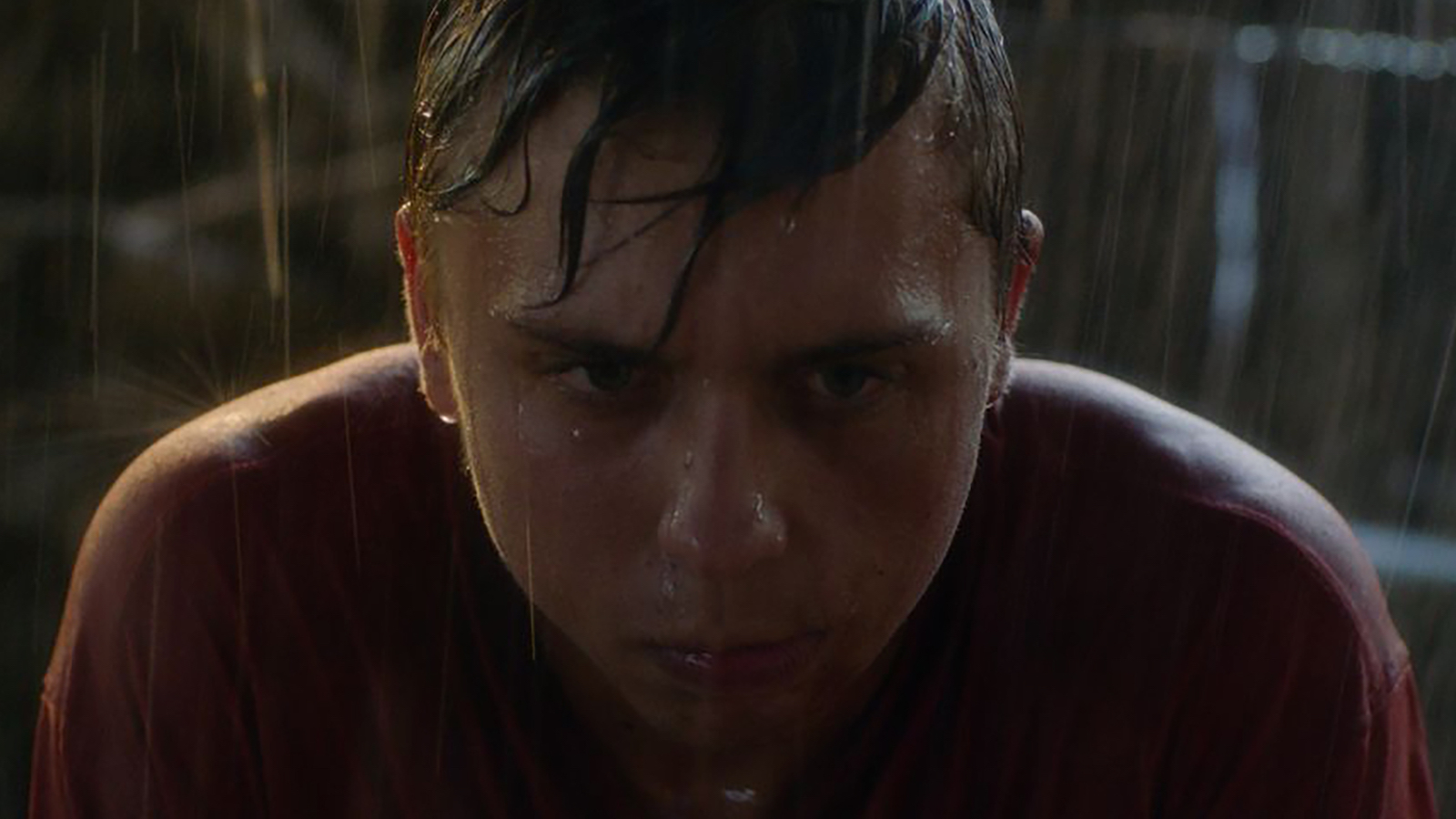 A boy in close-up with an intense face staring straight ahead covered in pouring rain