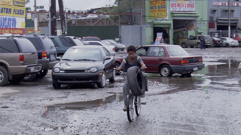 A young boy rides a bicycle through a wet parking lot with a car tire hanging from his handlebars