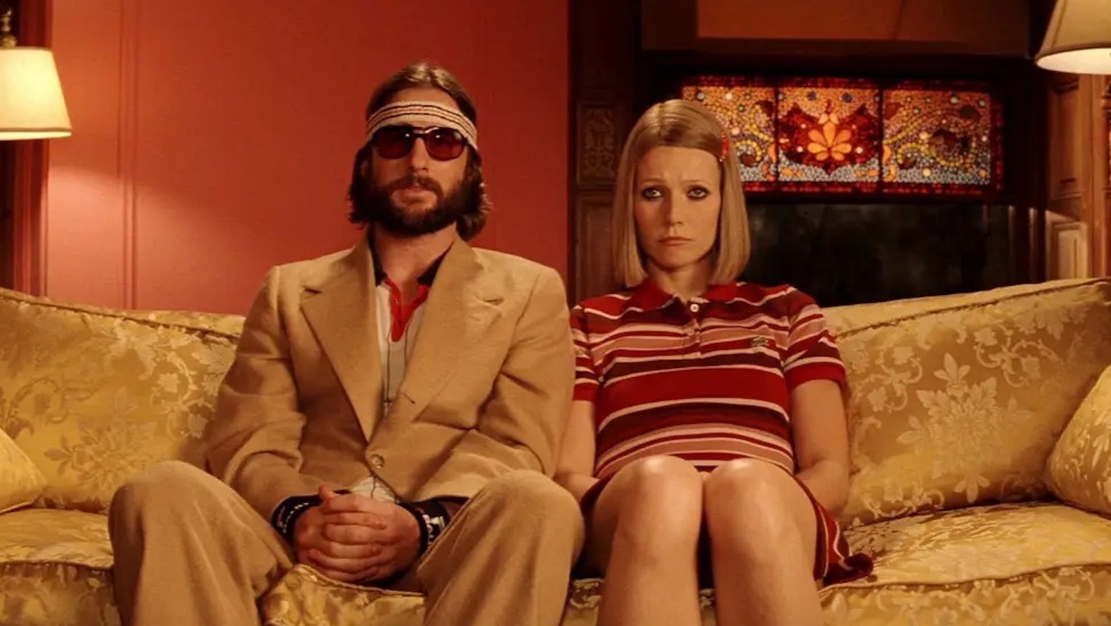 A man in a suit and bandana and a woman with medium length blond hair look morose and sit side by side on a couch looking at camera