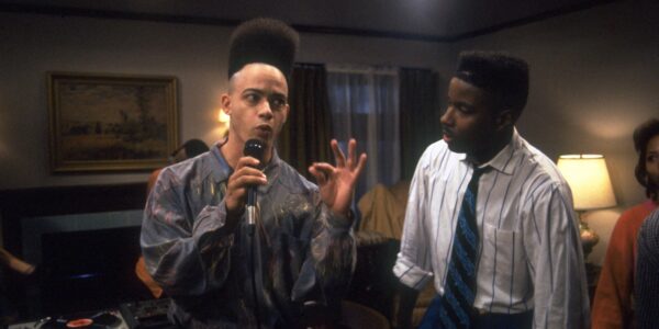 A man with a tall afro hairdo stands in a living room talking into a microphone while another man, wearing a shirt and tie, looks at him curiously