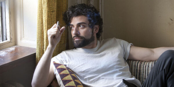 A man in a white t shirt and holding a cigarette looks out a window pensively