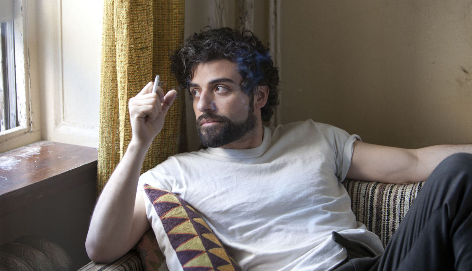 A man in a white t shirt and holding a cigarette looks out a window pensively