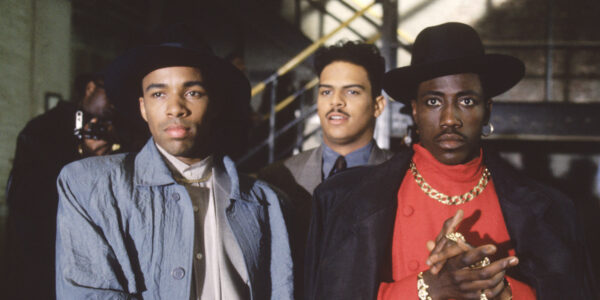 Two men in fedoras, one wearing a red shirt and gold chain necklace and rubbing his hands together look at camera, with a third man in a suit standing behind them