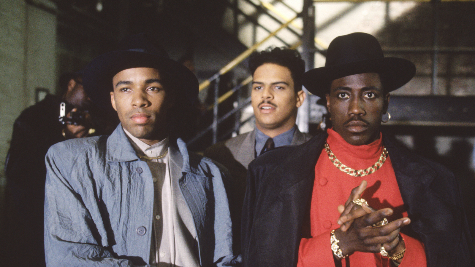 Two men in fedoras, one wearing a red shirt and gold chain necklace and rubbing his hands together look at camera, with a third man in a suit standing behind them