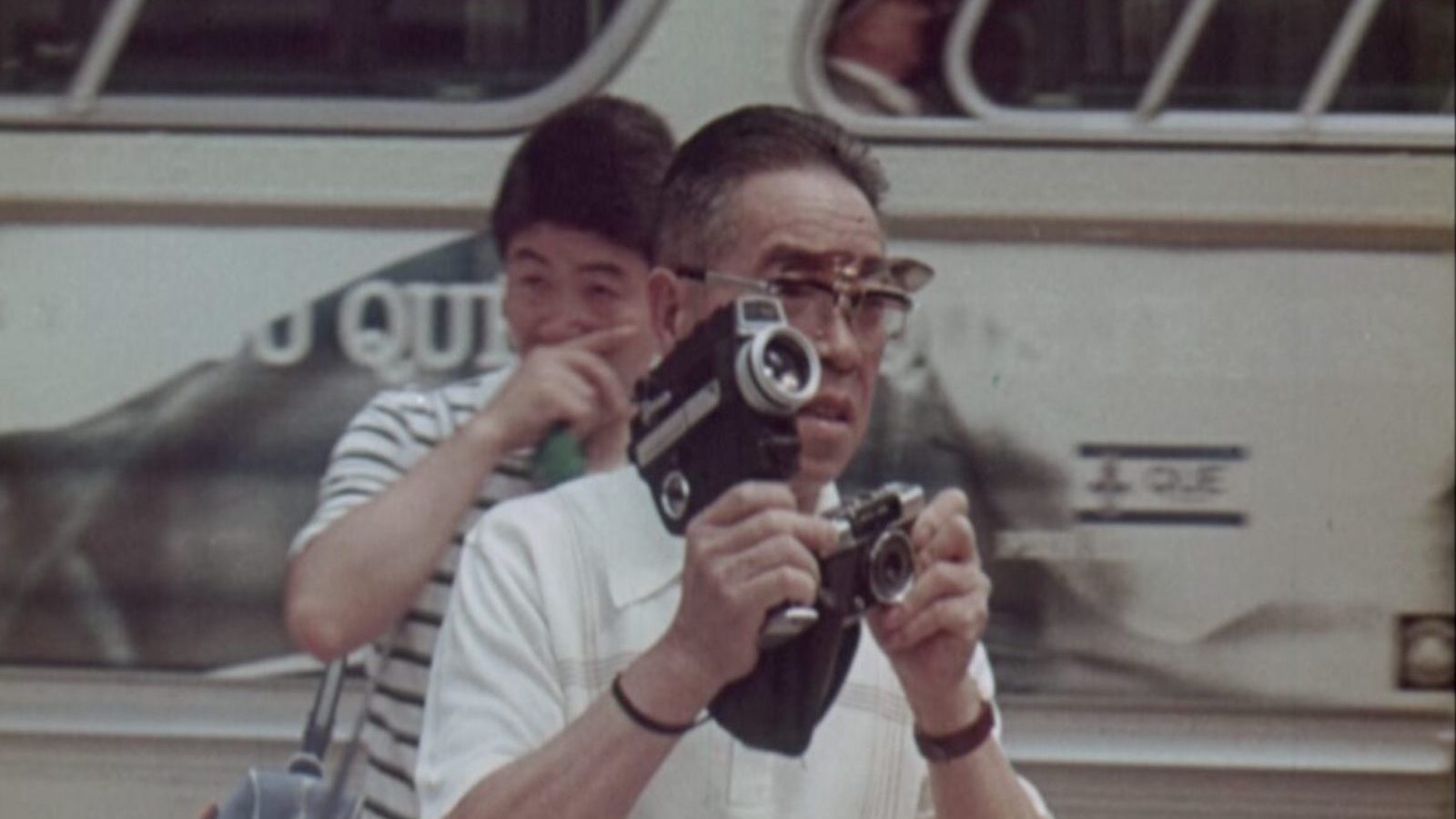 A man with flip sunglasses holds an 8mm camera and a photography camera simultaneously as he stands in front of the side of a bus, with a young man in a striped shirt behind him