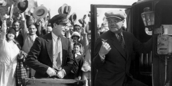 A man in glasses and a suitcase holds a suitcase next to a man in a chapeau getting in a car in a black-and-white photo
