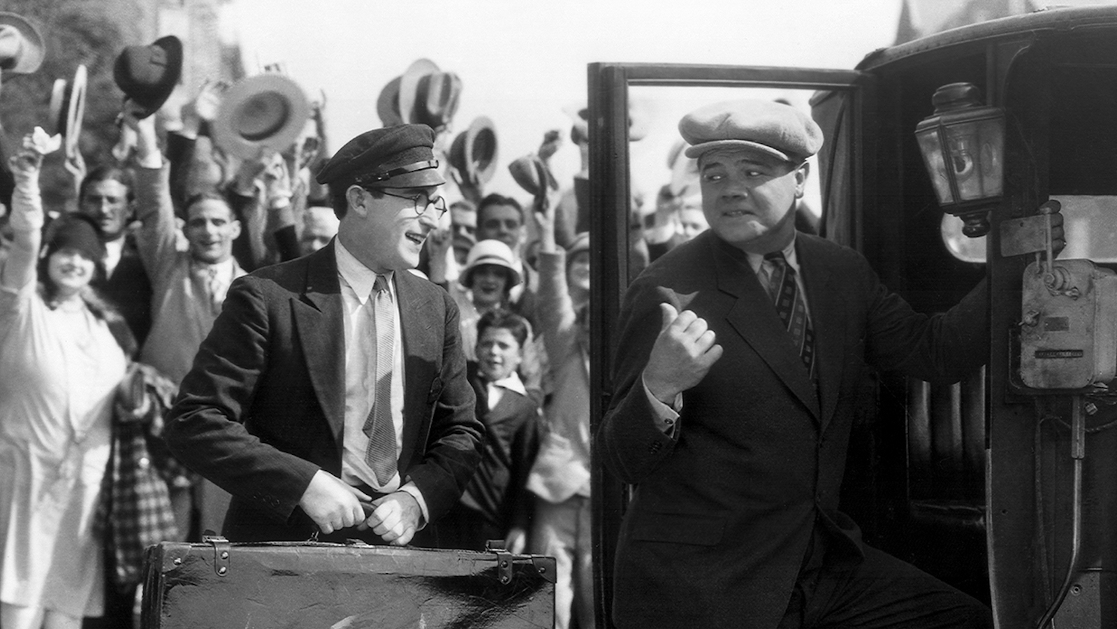 A man in glasses and a suitcase holds a suitcase next to a man in a chapeau getting in a car in a black-and-white photo