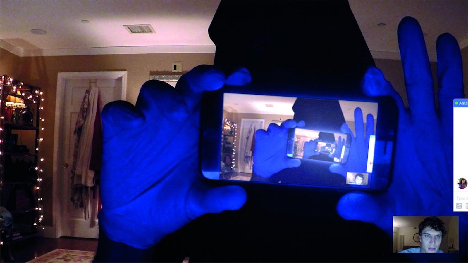 A hooded figure holds a cell phone that shows the same image over and over in a hall of mirrors