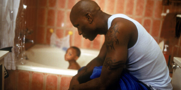 A man in a white tank top sits on a toilet, leaning over, with a young child, out of focus in the bathtub behind him