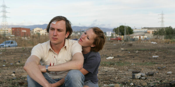 A man with a bandage on his head is embraced by a woman while sitting in a dump lot