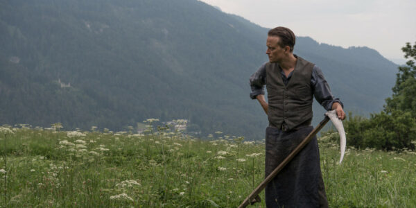 A man holds a scythe in a field of grass amidst mountains