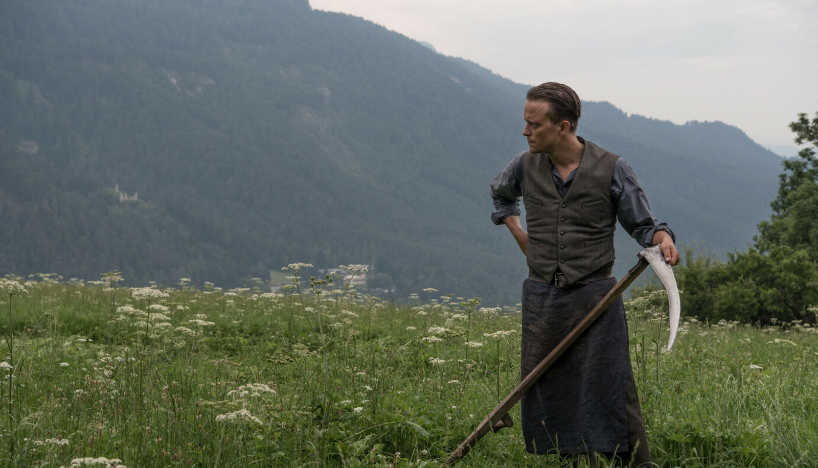 hiddenlife A man holds a scythe in a field of grass amidst mountains