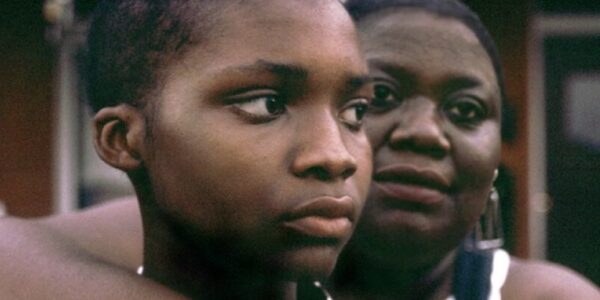 A mother holds her young son with her arm around him from the back, they both face camera