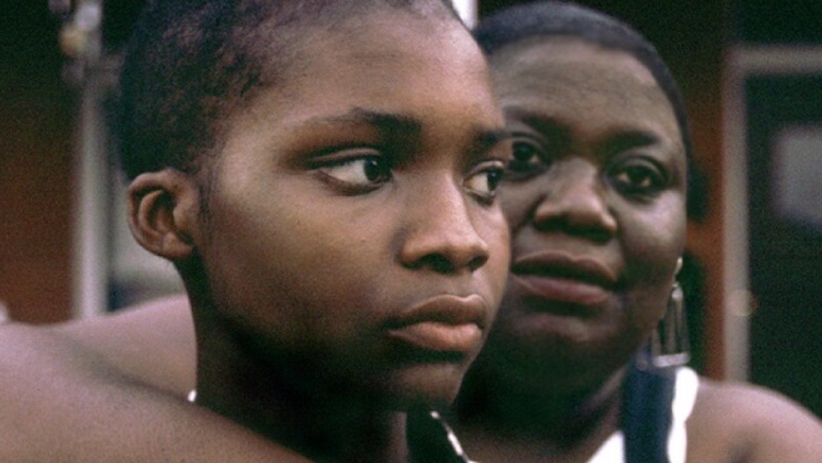 A mother holds her young son with her arm around him from the back, they both face camera