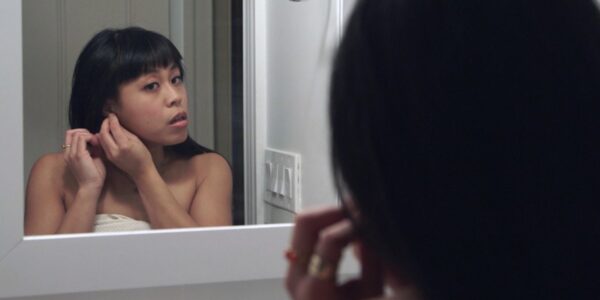 A woman with long hair puts on earrings looking in a mirror