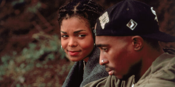 A young woman and man sit outside, she's behind him, glancing his way sympathetically while the man, wearing a backwards baseball cap in the foreground and looking offscreen