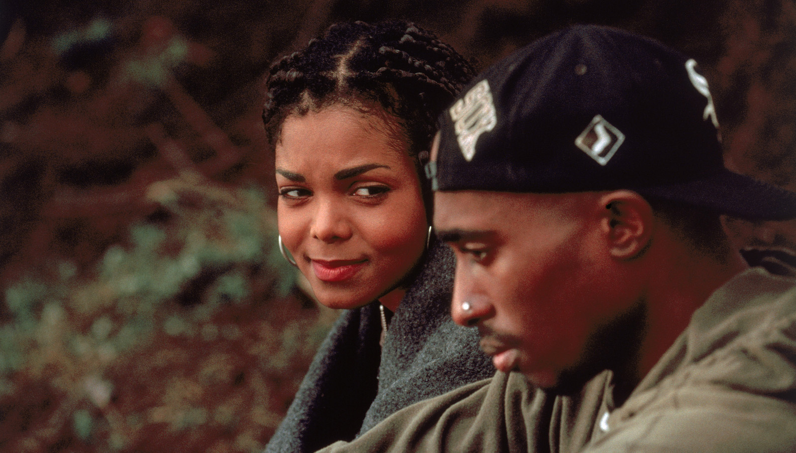 A young woman and man sit outside, she's behind him, glancing his way sympathetically while the man, wearing a backwards baseball cap in the foreground and looking offscreen