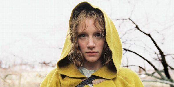 A young woman in a yellow hooded jacket looks at camera ominously in front of bare tree branches