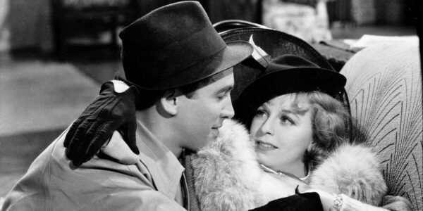 A man in a stetson hat and a woman with a feather in her hat embrace on a couch in a black and white image