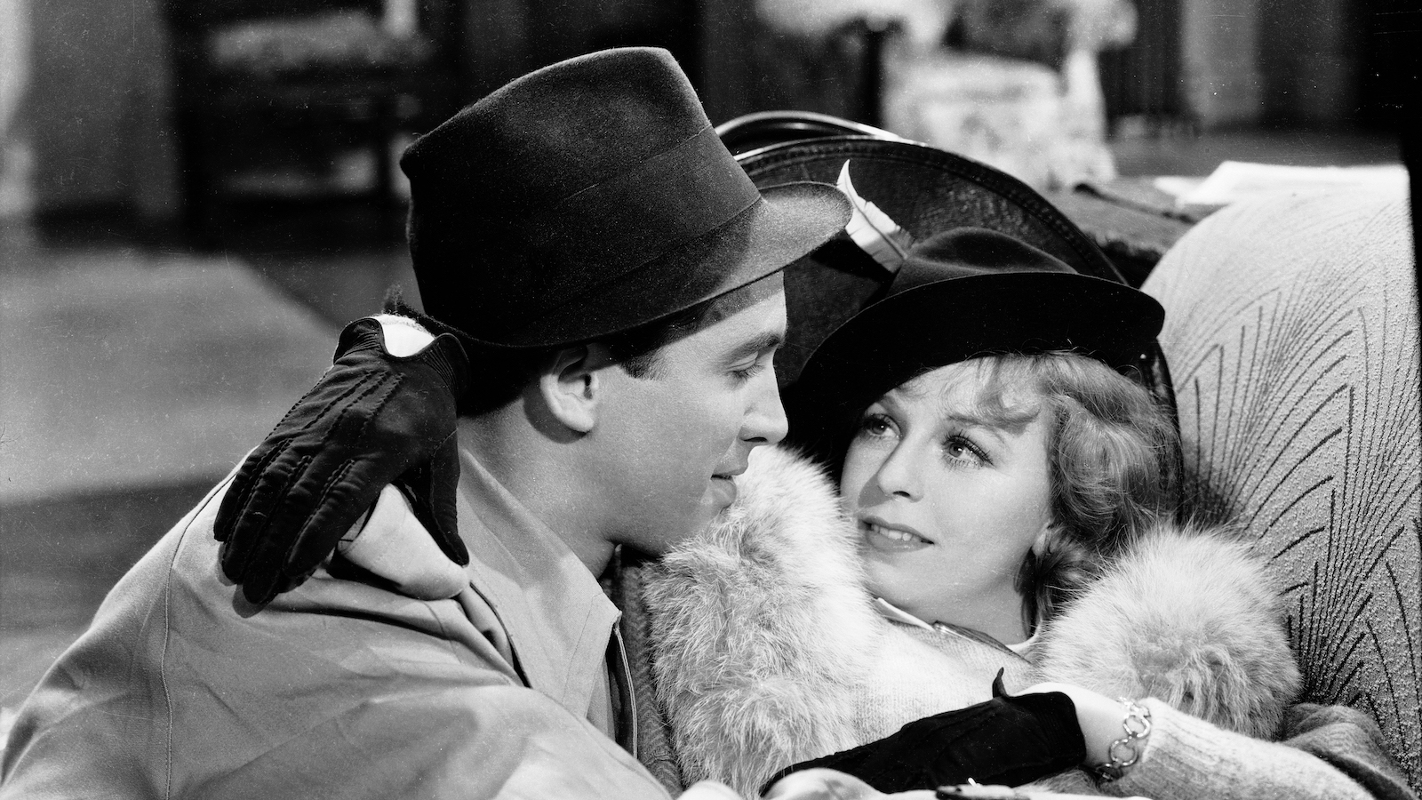 A man in a stetson hat and a woman with a feather in her hat embrace on a couch in a black and white image
