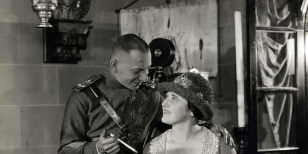 A man in military uniform holding a long cigarette looks down and smiles at a woman in a hat he stands over