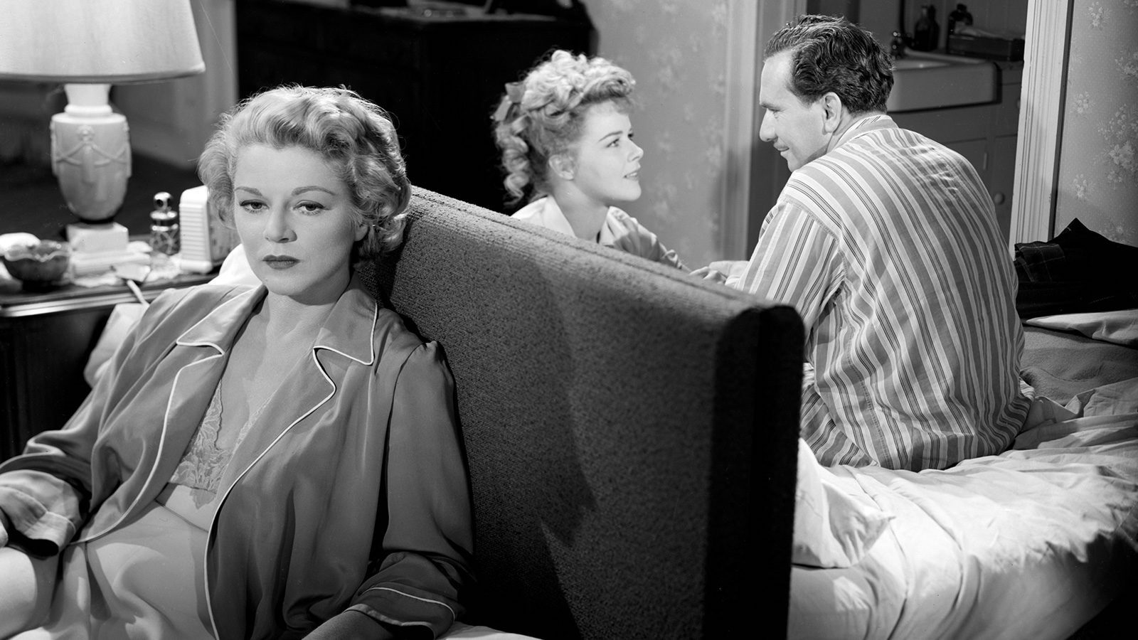 A woman sits on a couch, listening intently to an older man and a young woman behind her, her face turned to camera and away from them