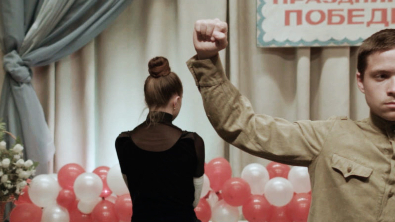 A woman faces away from the camera in a room set for a celebration while a man in a soldier's uniform raises his arm
