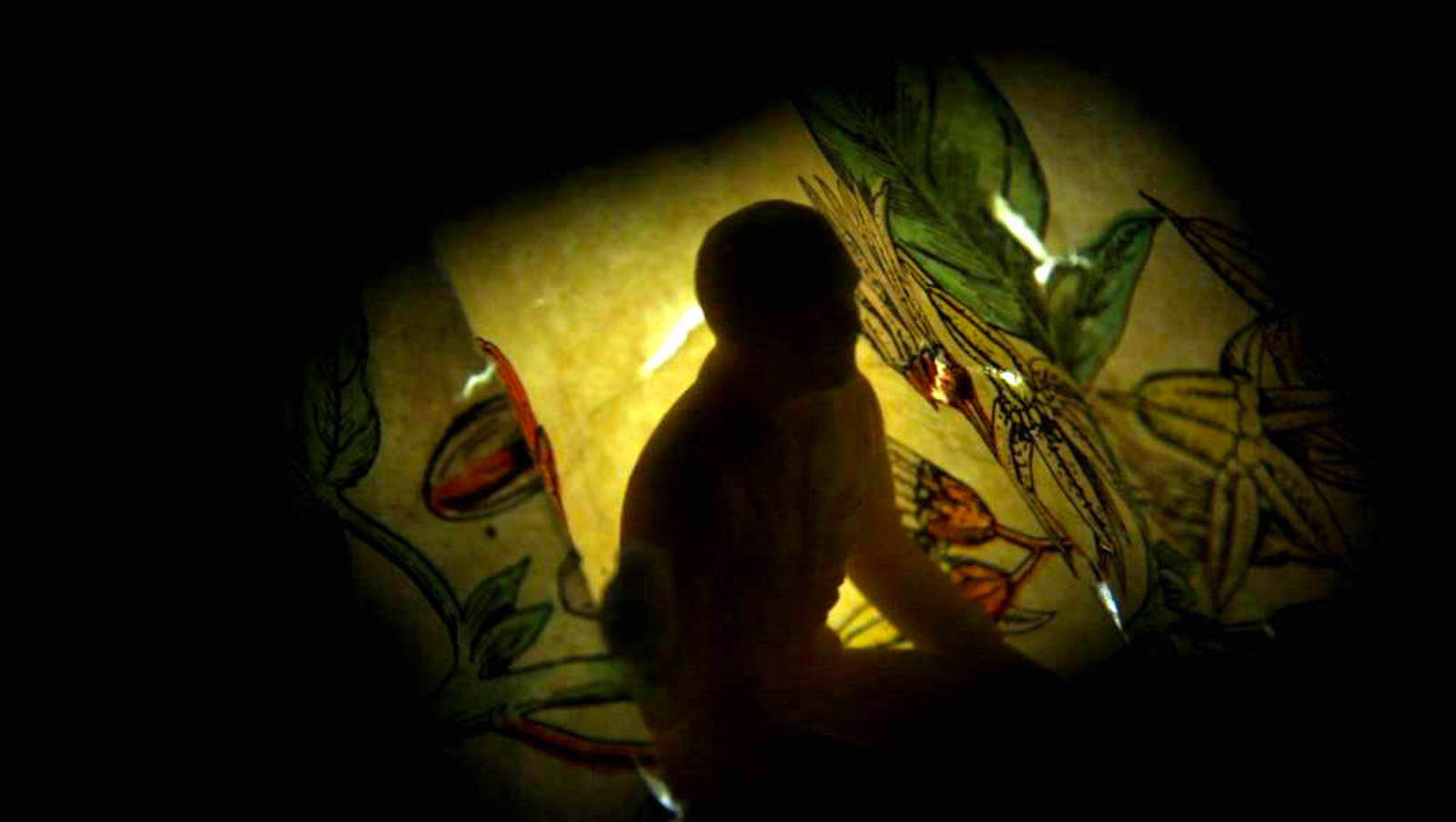 A figure of a man in silhouette hunched over in front of yellow paper walls with light shining through