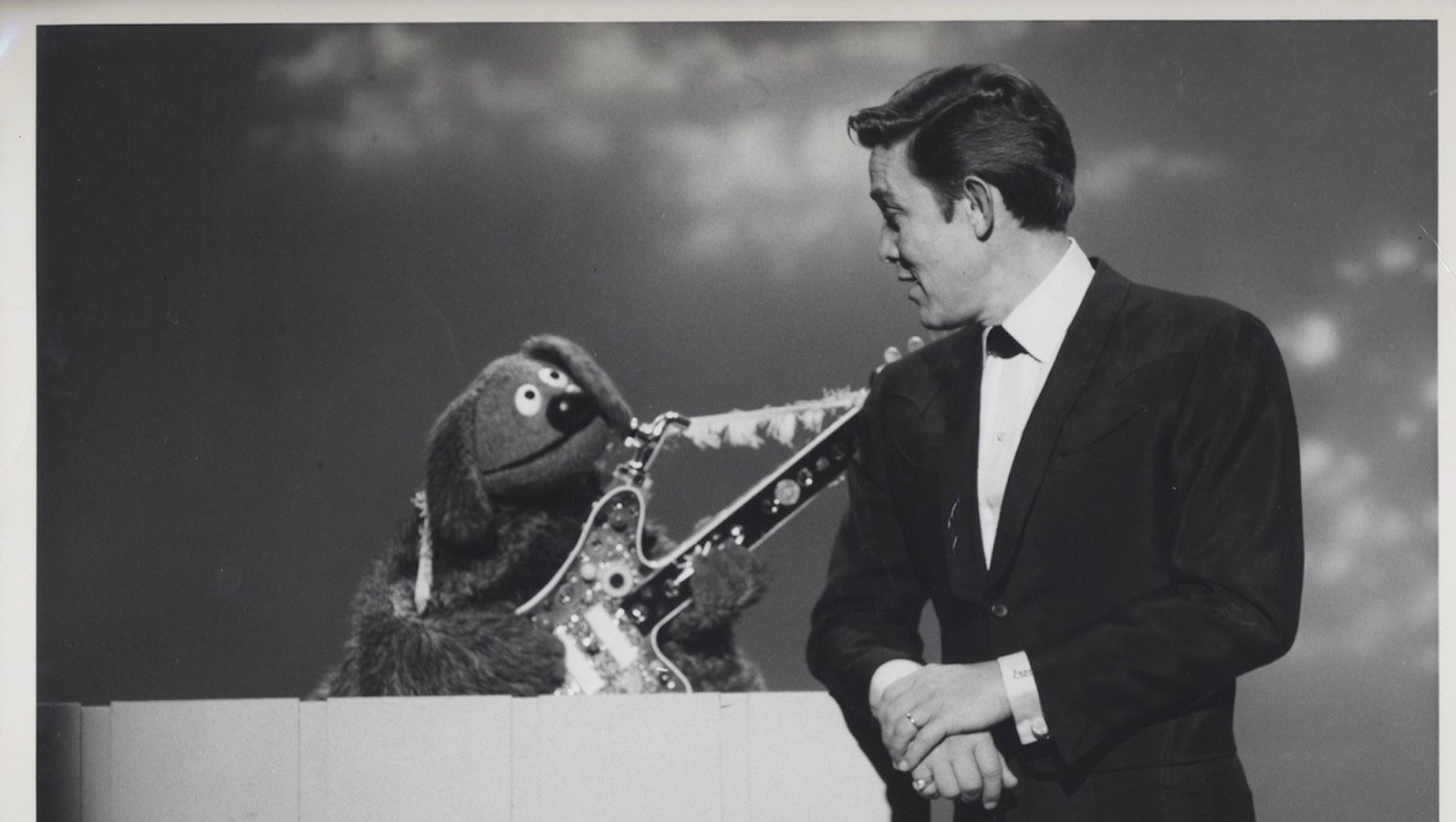 The dog muppet Rowlf holds a guitar and looks up at a man in a suit in a black-and-white image.