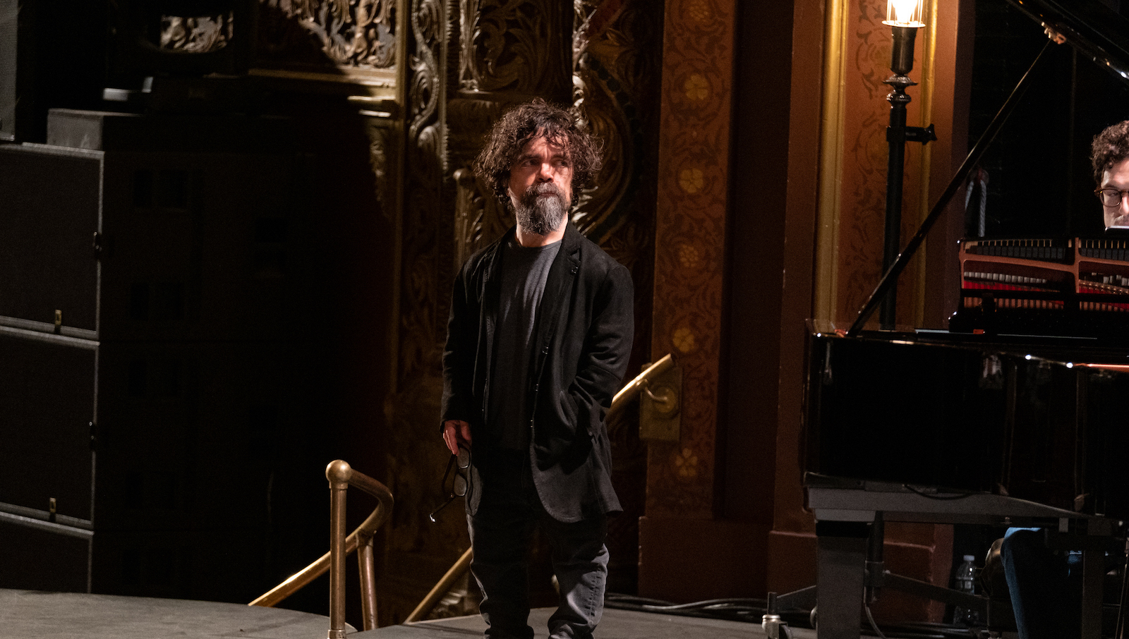 A little person wearing a beard stands backstage at a theater, one hand in his pocket, his hair curly and messy.