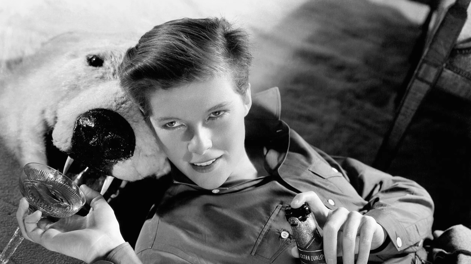 A young woman with a boy's haircut reclines on a stuffed bear's head on the floor and holds a champagne bottle in one hand and a champagne glass in the other, smiling past the camera