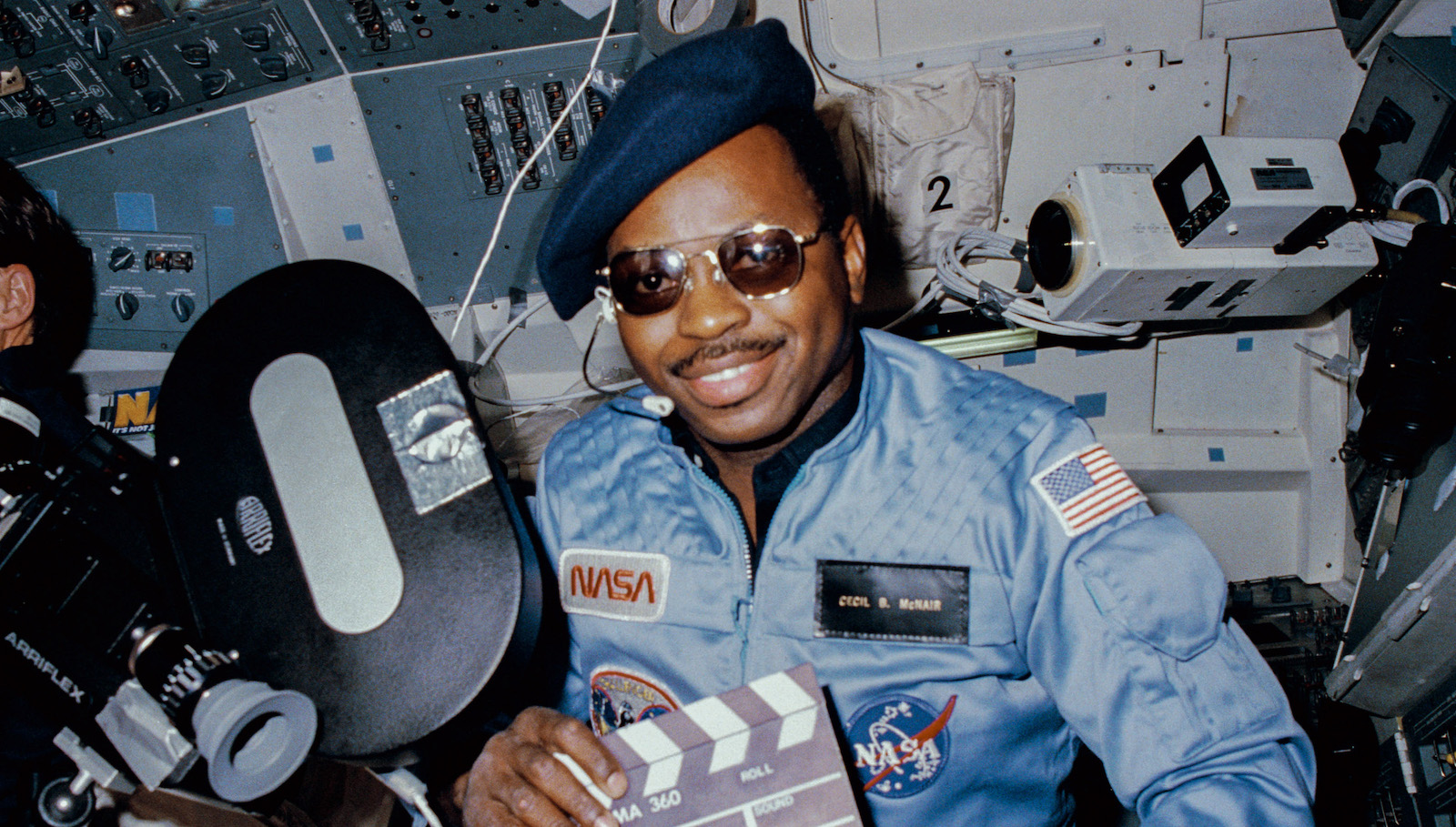 A man in hat and sunglasses and holding a film slate sits in a space shuttle smiling at camera